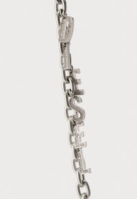 Srebrna metalna narukvica s lancem i riječi "DIESEL" u blok slovima, ukrašena prozirnim cirkonima na teksturiranoj površini.