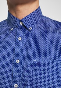 Camicia button-up blu con un piccolo motivo a quadretti bianchi, dotata di un colletto classico, bottoni bianchi e un logo sottile sulla tasca.