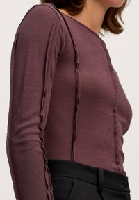 Langsleeve, geribbelde top in diep bordeaux met een ronde hals. Kenmerkt zich door golvende naadtoevoegingen en wordt gecombineerd met zwarte, strakke broeken.