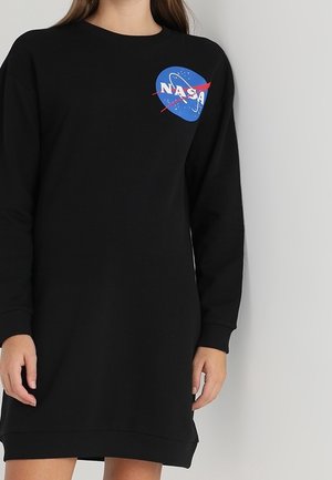 Frau trägt ein schwarzes, langärmeliges Kleid mit einem blauen NASA-Logo auf der linken Brust vor einem schlichten hellgrauen Hintergrund.
