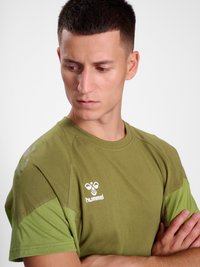 Jeune homme aux cheveux courts portant un t-shirt Hummel vert olive regarde vers le bas sur sa gauche, les bras croisés, sur un fond blanc.