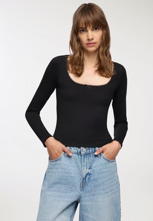 Top nero a maniche lunghe con scollatura a barca e bordo in pizzo, abbinato a jeans a vita alta di colore blu chiaro. Tessuto liscio con una vestibilità aderente.