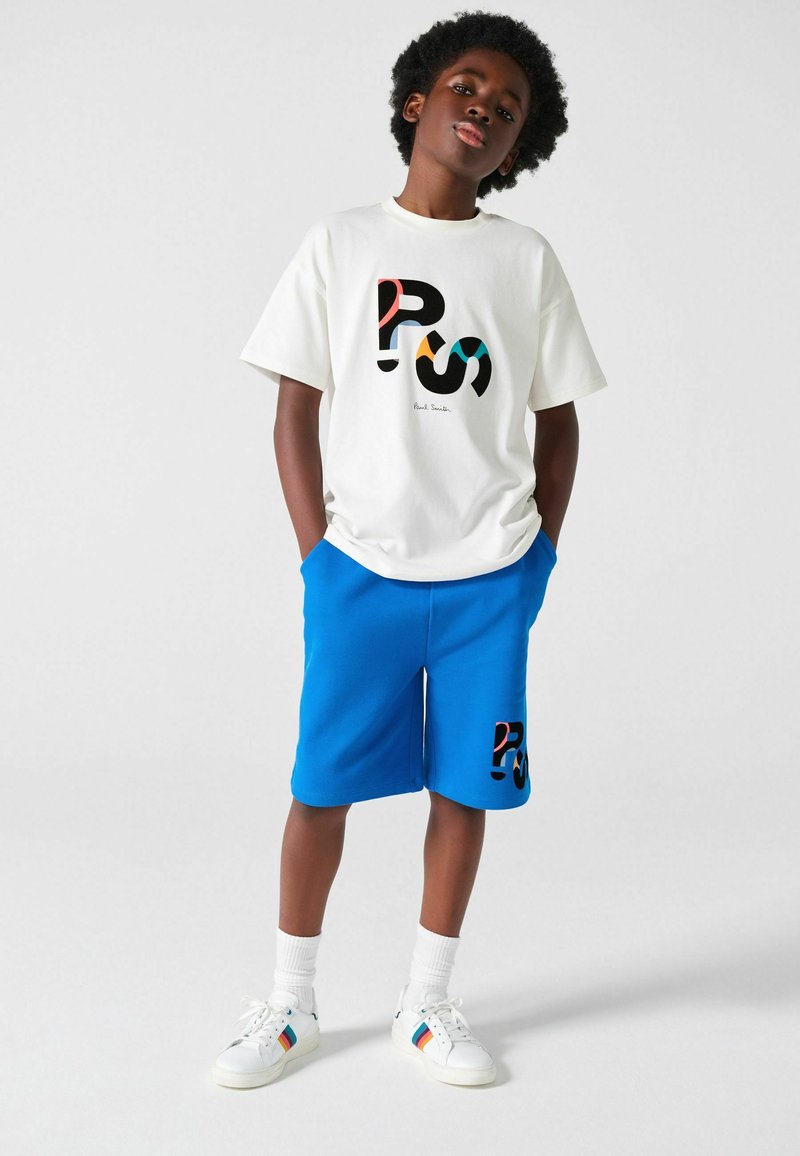 Chico de pie con las manos en los bolsillos, lleva una camiseta blanca con gráfico, pantalones cortos azules con el mismo logo, calcetines blancos y zapatillas blancas con rayas de colores.