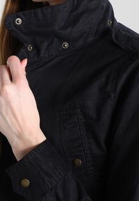 Černá, lehká bunda s vysokým límcem, patentním zapínáním a strukturovanou tkaninou. Viditelná náprsní kapsa a kovové detaily.