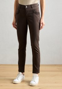 Hnedé slim-fit nohavice vyrobené z bavlneného zmesi, s piatimi vreckami, zapínaním na gombík a hladkým povrchom, skombinované s bielymi teniskami.