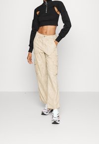 Czarny krótki sweter z wycięciami i detalem na zamek, sparowany z beżowymi spodniami cargo w wzory. Model ma na sobie białe i czerwone sneakersy.