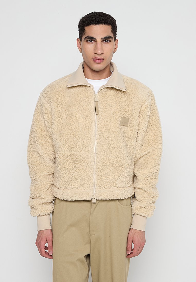 Chaqueta de forro polar beige con cremallera frontal y cuello, que presenta puños y dobladillo acanalados, y un pequeño parche con el logo en el lado izquierdo del pecho.