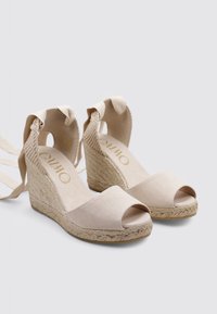 Alpargatas beige con cuña, punta abierta, parte superior de tela, suela de yute trenzado y lazos de cinta alrededor del tobillo. Textura suave y diseño minimalista.