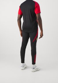 Schwarzes und rotes Sportoutfit mit einem kurzärmeligen Oberteil und taillierten Hosen mit roten Streifen. Der Stoff ist glatt und eng anliegend. Silberne Sportschuhe.