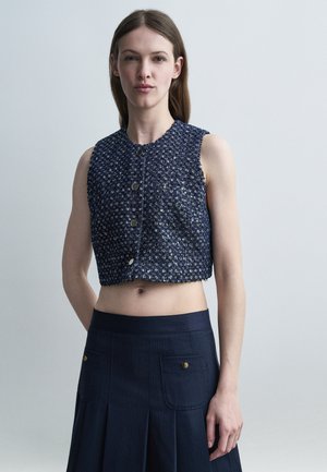 Νεαρή γυναίκα με ίσια μαλλιά φορά αμάνικο cropped γιλέκο σε ναυτικό μπλε με υφή και μεταλλικά κουμπιά, καθώς και φούστα ψηλόμεση σε ναυτικό μπλε με μπροστινές τσέπες.