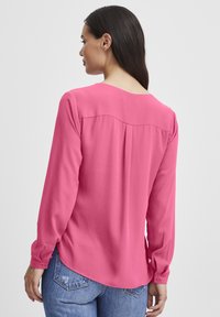 Pink long-sleeve blouse made of smooth fabric, featuring a round neckline, back pleat detail, and a curved hem. Worn with blue jeans.