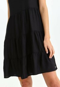 Schwarz ärmelloses Kleid mit gestuften Rüschen, aus leichtem Stoff. Besitzt ein subtiles Logo-Accessoire nahe dem Saum. Glatte Textur.