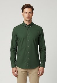 Camisa verde de manga larga con botones y cuello, hecha de una tela suave. Presenta botones blancos y un corte ajustado. Combinada con pantalones color claro.