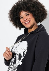Zwarte hoodie met een grote witte schedelafbeelding, versierd met metalen accenten en een zilveren trekkoord. Zachte stoftextuur.