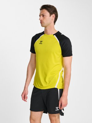 Mężczyzna ubrany w żółto-czarną sportową koszulkę z krótkim rękawem i czarne szorty z białymi detalami, stojący i patrzący na bok na białym tle.