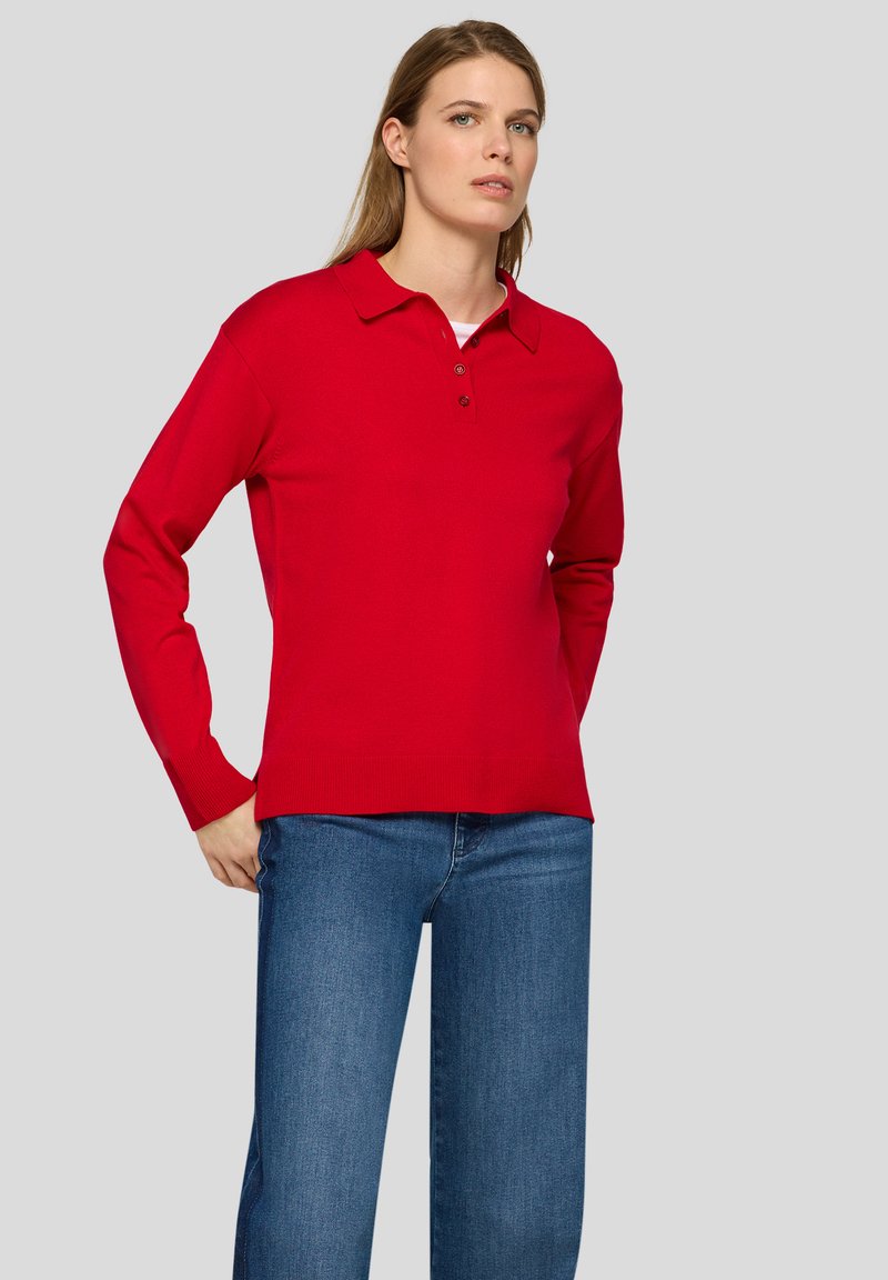 Rot gestrickter Pullover mit Polo-Kragen, drei Knöpfen, langen Ärmeln und geripptem Saum. Kombiniert mit blauen Jeans.