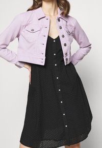 Chaqueta de mezclilla lavanda corta con detalles de botones plateados, bolsillos y dobladillo deshilachado; llevada sobre un vestido negro de botones texturizado.