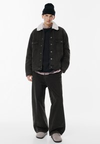 Giubbotto di jeans nero con colletto di pelliccia bianca, indossato sopra un maglione blu marino e una camicia a quadri, abbinato a pantaloni neri larghi e scarpe grigie.
