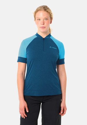 Frau mit hellen lockigen Haaren trägt ein kurzärmeliges blaues Radtrikot mit Frontreißverschluss und schwarze Hosen, stehend vor einem schlichten Hintergrund.