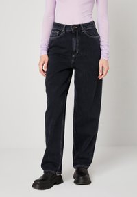 Sieviete, kas valkā augsta vidukļa brīvas melnas džinsas, melnas masīvas zābakus un gaiši violetu garroku blūzi, stāvot pret vienveidīgu gaišu fonu.