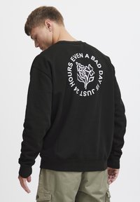 Svart hettegenser med rund hals og lange ermer. Baksiden har hvitt blomstermotiv og tekst. Materialet ser mykt ut med en avslappet passform.