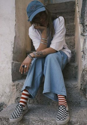 Buty wsuwane w kratę, szerokie dżinsowe spodnie, biała koszulka, niebieska czapka i czerwono-białe paskowane skarpetki. Stylowa biżuteria na warstwach.