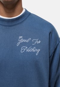 Felpa blu con colletto a coste, caratterizzata da ricami bianchi in corsivo che recitano "Good For Nothing" sulla parte anteriore. Tessuto morbido.