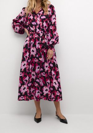 Femme portant une robe midi à manches longues boutonnée, avec un motif floral abstrait rose, violet et noir, et des chaussures noires à bouts pointus.