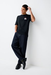 Črna majica s kratkimi rokavi z majhnim barvitim potiskom, v kombinaciji s temnimi denim hlačami in črnimi čevlji z debelim podplatom.