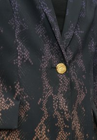 Blazer en tissu noir avec un motif serpent violet et beige, agrémenté d'un bouton doré texturé en guise d'accent.