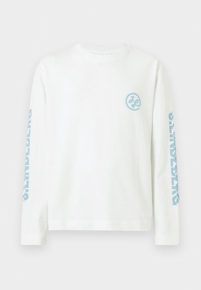 Λευκό πουκάμισο με μακριά μανίκια, με ανοιχτό μπλε κυκλικό λογότυπο στο αριστερό στήθος και τη λέξη «ORIGINALS» τυπωμένη κατακόρυφα σε ανοιχτό μπλε χρώμα και στα δύο μανίκια.