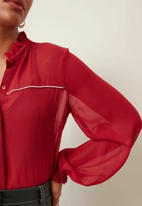 Blusa roja semitransparente con un cuello con volantes, mangas largas y un ribete decorativo blanco en el pecho. Presenta cierres de botones y puños fruncidos.