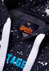 Granatowy hoodie z białymi sznurkami i wzorem w plamy farby, z pomarańczową i białą metką marki SuperDry umieszczoną wewnątrz kołnierza.