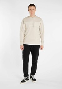 Beige sweatshirt met NXG-logo, ronde hals, en geribde manchetten, gecombineerd met zwarte broeken en zwarte hoge sneakers. Gladde textuur.