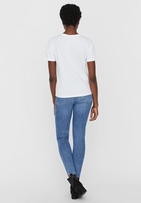 Witte katoenen t-shirt met korte mouwen, gecombineerd met blauwe skinny jeans van denim. Model draagt zwarte enkelhoge laarzen. Effen achtergrond.