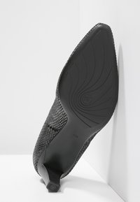 Chaussure noire à talon haut avec un dessus texturé, un bout pointu et une semelle en caoutchouc lisse présentant un motif en vagues.