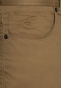 Khaki spodnie uszyte z fakturowanej tkaniny, z naszywaną kieszenią i wytłoczonym logo na kieszeni. Zawiera szczegół z czarnym metalowym guzikiem.