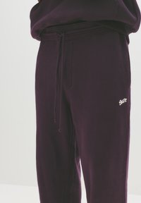 Pantaloni de trening violet, cu un umăr ajustabil și un mic logo alb. Material moale, cu o croială lejeră și detalii subtile de textură.