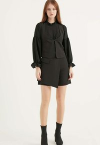 Zwarte outfit bestaande uit een aangesloten vest met ritssluiting, lange pofmouwen en shorts tot de knie. Gladde stof met gerimpelde accenten bij de halslijn.