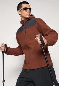 Veste en polaire marron avec un panneau d'épaule noir, poches zippées et surface texturée. Le modèle tient des bâtons de trekking et porte des lunettes de soleil.