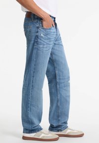 Heledad sinine teksapüksid, millel on mugav lõige, kulumis- ja kahjustuskohtadega, küljetaskud ning pruun nahkplekk vöökoha juures.