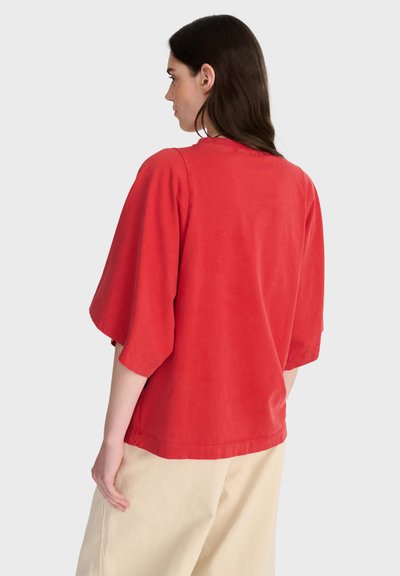 Femme aux longs cheveux foncés portant un haut rouge ample à manches larges et un pantalon beige clair, vue de dos sur un fond uni.