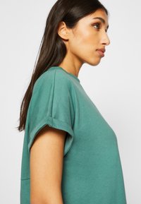 Teal bomull t-skjorte med rund hals, rullet kort erm, avslappet passform og jevn tekstur, vist på en modell fra siden.