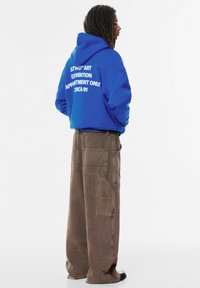 Felicità blu royal con testo bianco sulla schiena; abbinato a pantaloni cargo marroni larghi con molte tasche e una vestibilità rilassata.