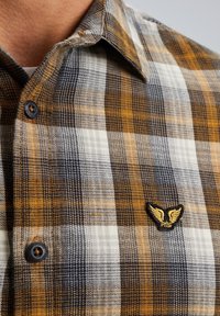 Braun-, Weiß- und Grau-kariertes Flanellhemd mit einem schwarzen Knopf und einem gestickten Logo mit goldenen Flügeln auf der Brust.