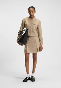 Beige striped shirt with two front pockets and a pleated mini skirt. Model holds a black triangular bag. Black shoes with white socks.