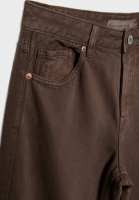 Jean en denim marron montrant la poche avant, les passants de ceinture, le bouton et l'étiquette de taille à l'intérieur de la ceinture.