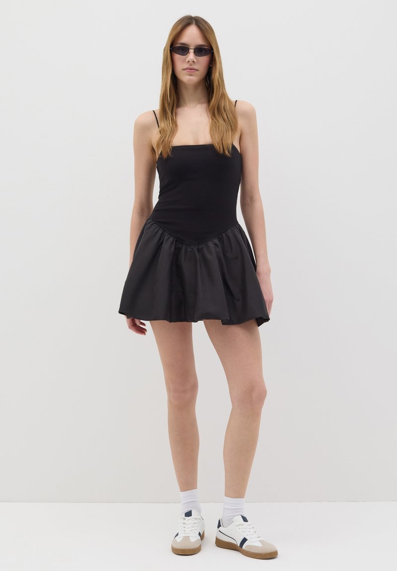 Robe noire mini avec un haut ajusté et une jupe en satin évasée. Le mannequin porte des lunettes de soleil noires élégantes et des baskets blanches avec des accents marins.