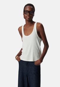 Witte geribde tanktop met een ronde halslijn, gecombineerd met donkerblauwe wijde jeans. Model staat met handen in de zakken en draagt een bril met zwarte montuur.