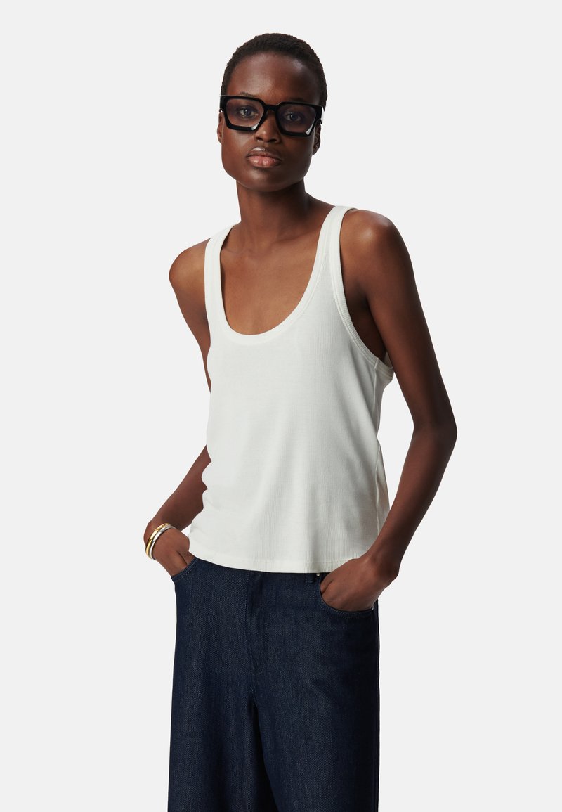 Witte geribde tanktop met een ronde halslijn, gecombineerd met donkerblauwe wijde jeans. Model staat met handen in de zakken en draagt een bril met zwarte montuur.